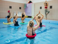 Eine Masseurin und medizinische Bademeisterin  leitet eine Gruppe von Frauen im Wasser an, die gemeinsam Yoga-Übungen im Schwimmbecken durchführen.