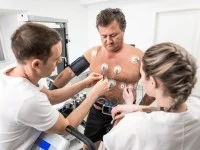 Eine medizinische Technologin und ein Technologe für Funktionsdiagnostik befestigen Messelektroden am nackten Oberkörper eines Patienten. 