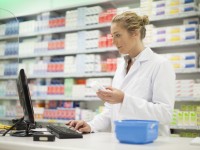 Eine pharmazeutisch-technische Assistentin im weißen Laborkittel recherchiert am Computer den Bestand eines Hautpflegeprodukts.