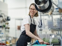 Eine Assistentin für Ernährung und Versorgung putzt die Küchenspüle in einem Heim für Menschen mit Behinderungen. Dabei trägt sie einen schwarzen Kittel und eine Brille.