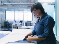 Eine bekleidungstechnische Assistentin schneidet auf einem Tisch in einer hellen Halle eine Entwurfsskizze zu einen Musterschnitt zurecht. 