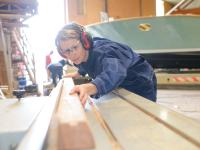Eine Bootsbauerin prüft in ihrer Werkstatt ein geschnittenes Holzstück.