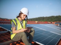 Eine technische Assistentin für regenerative Energietechnik sitzt  in Sicherheitsausrüstung mit einem Tablet in der Hand auf dem Dach neben den PV-Panelen.