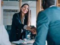 Eine Frau im Businessoutfit begrüßt einen ausländischen Mann per Handschlag. Ein weiterer Mann beobachtet das Geschehen.