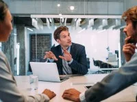 Ein Mann im Business-Outfit berät in Pärchen im Business-Outfit. Alle sitzen an einem Tisch, auf dem ein Laptop und Wasser steht. 