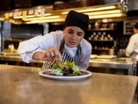 Eine gastronomische Assistentin garniert eine Speise in der Großküche. Im Hintergrund arbeitet eine weitere Person aus dem Betrieb.