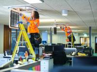 Eine Elektronikerin für Gebäude- und Infrastruktursysteme installiert mit ihrem Kollegen eine Klimaanlage in einem Bürogebäude.