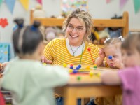 Eine lächelnde sozialpädagogische Assistentin / Kinderpflegerin spielt mit vier Kleinkindern am Tisch in einem Kinderhort.