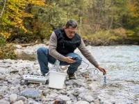 Ein umweltschutztechnischer Assistent hockt neben einem Flusslauf und hält ein Messgerät in das Wasser.