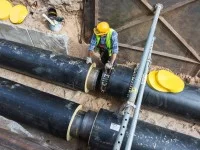 Ein Leitungsbauer steht in einer Grube, in der zwei große Rohre verlaufen. Er trägt Sicherheitskleidung und stützt sich an einem Rohr ab. 