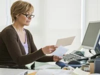 Eine kaufmännische Assistentin / Wirtschaftsassistentin für Büro und Sekretariat hält zwei Briefe in der Hand, während sie vor ihrem Computer im Büro sitzt.