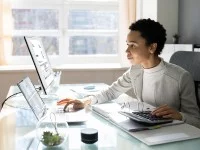 Eine kaufmännische Assistentin / Wirtschaftsassistentin für Fremdsprachen sitzt an ihrem Schreibtisch im Büro und tippt Zahlen, die sie mit Hilfe eines Taschenrechners berechnet, in ihren Laptop. 