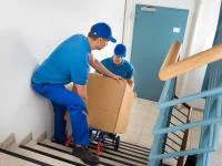 Zwei Fachkräfte für Möbel-, Küchen- und Umzugsservice transportieren einen Umzugskarton mit einer Sackkarre die Treppe hinunter.