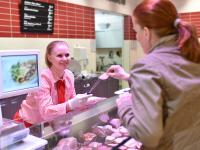 Eine Fachverkäuferin im Lebensmittelhandwerk bietet einer Kundin an der Fleischtheke ein Stück Wurst zum Probieren an.
