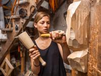 Eine Holzbildhauerin bearbeitet eine Skulptur mit Knüpfel und Beitel.