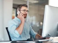 Ein kaufmännischer Assistent / Wirtschaftsassistent für E-Business Management sitzt an seinem Schreibtisch und telefoniert, während er auf seinen Computerbildschirm schaut.