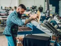 Ein Medientechnologe Siebdruck bedient eine Siebdruckmaschine zum Bedrucken von T-Shirts.