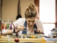 Eine Gold- und Silberschmiedin sitzt in ihrer Werkstatt an einem Tisch und schweißt zwei Glieder der neuen Kette zusammen.